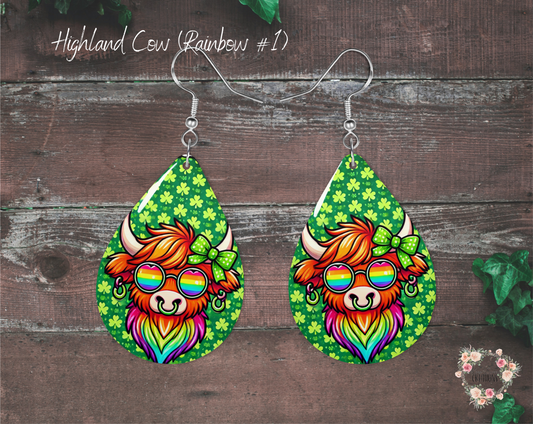 "A pair of teardrop-shaped St. Patrick’s Day earrings featuring a colorful Highland cow wearing rainbow sunglasses and a green polka-dot bow. The background is green with shamrocks, and the earrings are made from lightweight sublimation wood with nickel-free hypoallergenic hooks."