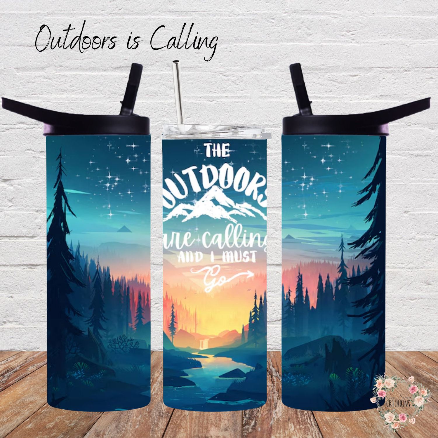 A stainless steel tumbler with a scenic outdoor design, featuring a mountain sunset, tall pine trees, and a reflection on a calm lake. The tumbler displays the phrase "Outdoors is Calling" in elegant script. The set includes tumblers with both metal straws and black flip-top lids. The background varies, including wood textures and white brick walls, enhancing the nature-inspired aesthetic.